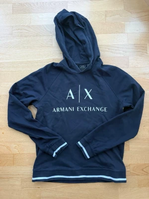 Marinblå hoodie från Armani Exchange - Marinblå hoodie från Armani Exchange med vit logga framtill. Hoodien har långa ärmar och ribbade muddar med vita detaljer vid ärmslut och nederkant. Tillverkad i mjukt bomullsmaterial och har en klassisk huva.