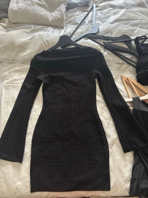 Svart långärmad kortklänning - Säljer en svart, figurnära kortklänning med lång ärm och lätt struktur i tyget. Klänningen har en enkel, stilren design med båtringning och passar perfekt för dig som gillar en clean look.