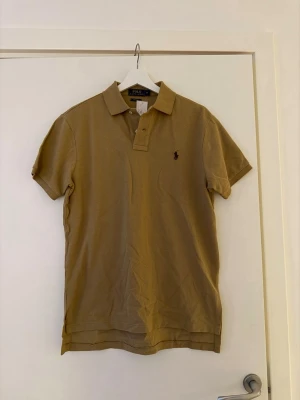 Beige pikétröja från Ralph Lauren - Säljer en beige pikétröja från Ralph Lauren i storlek M. Tröjan har klassisk krage, korta ärmar och två knappar vid halsen. Materialet är bomull och modellen är custom fit. Den har den broderade loggan på bröstet. Har aldrig använts 