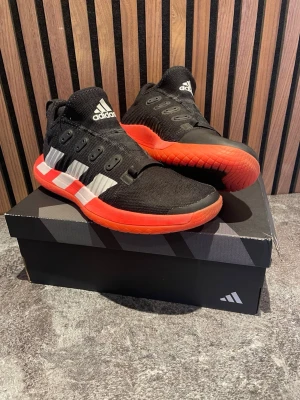 Svarta Adidas inneskor med röd/rosa sula - Snygga svarta inneskor från Adidas med tydliga vita ränder på sidan och en rosa/röd yttersula. Ovandelen är i textil för bra andningsförmåga och komfort. Klassisk snörning och logga på plösen. Perfekta för dig som gillar handboll.