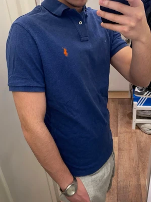 Polo Ralph Lauren (Blå piké) - Snygg blå piké från Polo Ralph Lauren med klassisk krage och knappar framtill. Ikonisk röd broderad logga på bröstet. Den är oanvänd och redo för en ny ägare! Pikén är storlek L men passar även M!