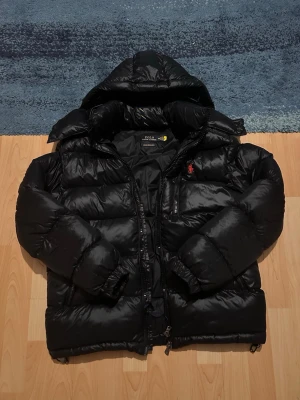 Svart pufferjacka från Polo Ralph Lauren - Säljer en svart pufferjacka från Polo Ralph Lauren med huva och dragkedja. Jackan har en röd logga på bröstet, flera fickor med dragkedja och är tillverkad i glansigt material. Perfekt för kalla vinterdagar och riktigt snygg streetstil.