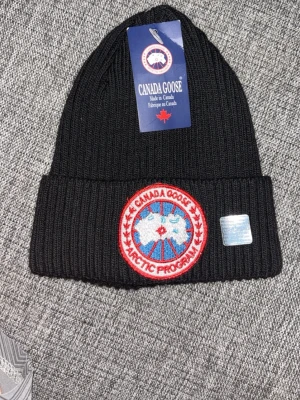 Svart ribbstickad mössa Canada Goose - Säljer en svart ribbstickad mössa från Canada Goose med klassisk uppvikt kant. Framtill sitter det ikoniska runda Arctic Program-märket i rött, vitt och blått samt en hologrametikett. Perfekt accessoar för kalla dagar.