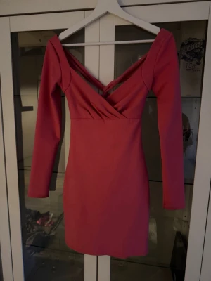 Rosa omlottklänning med v-ringning - Snygg rosa kortklänning med omlottdetalj och djup v-ringning både fram och bak. Klänningen har långa ärmar och en figurnära passform som framhäver siluetten. Perfekt för dig som vill sticka ut med en trendig och feminin look.