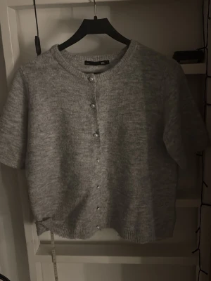 Grå kortärmad kofta med pärlknappar - Säljer en söt grå kofta med korta ärmar och pärlknappar framtill. Koftan är stickad i mjukt material och har rund halsringning samt ribbade kanter. Perfekt att styla över ett linne eller t-shirt för en chill look.