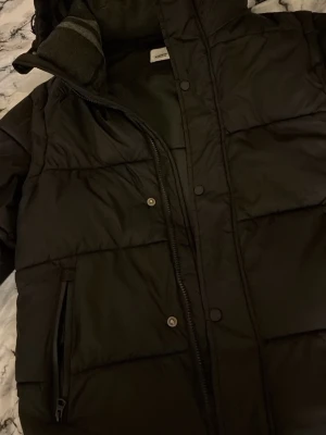 Svart pufferjacka från Burberry - Säljer en svart pufferjacka från Burberry med huva och dragkedja framtill. Jackan har quiltad design, sidofickor med dragkedja och en klassisk look. Perfekt för kalla dagar och riktigt snygg till vintern. Materialet är slitstarkt och håller värmen bra.