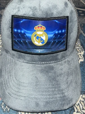 Grå Real Madrid keps med nät - Snygg grå keps med Real Madrid-logga framtill och blå bakgrund. Kepsen har böjd skärm och mesh på sidorna och bak för extra ventilation. Perfekt för dig som vill visa vilket lag du hejar på och samtidigt hålla stilen.