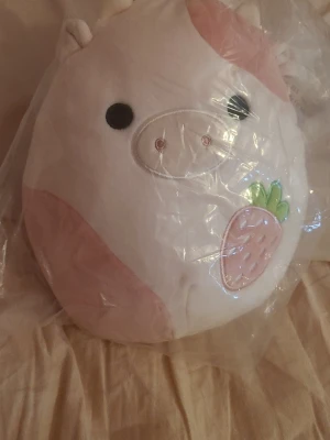 Squishmallow Reshma kudde rosa/vit - Supermjuk och rund Squishmallow-kudde i form av en söt ko med rosa och vita partier samt ett broderat jordgubbsmotiv. Modellen heter Reshma och har små horn och öron. Perfekt som mysig inredningsdetalj eller gosedjur.