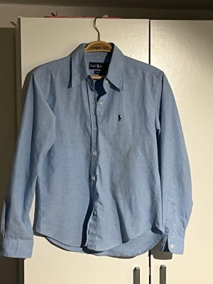 Ljusblå skjorta från Ralph Lauren - Klassisk ljusblå skjorta från Ralph Lauren med broderad logga på bröstet. Skjortan har lång ärm, knappar framtill och en tidlös krage. Tillverkad i mjuk bomull för en bekväm känsla och snygg passform.