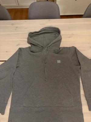 Grå hoodie från Acne Studios - Säljer en äkta stilren grå hoodie från Acne Studios med det klassiska Face-logotypen på bröstet. Hoodien har luva och en stor magficka. Tillverkad i mjuk bomullsblandning och har en relaxed fit som passar perfekt till streetwear. Säljer då den är lite för kort för mig, ungefär 180, pris går att diskutera vid snabb affär! Hör gärna av er!