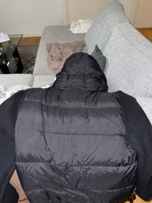 Svart cardigan från Moncler - Säljer en svart pufferjacka från Moncler med stickade ärmar och huva. Jackan har dragkedja framtill, två fickor med dragkedja och Moncler-logga på ena ärmen. Perfekt för kalla vinterdagar och riktigt snygg streetstil.