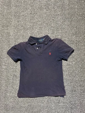 Mörkblå piké från Polo Ralph Lauren - Hej! Säljer denna Klassiska mörkblå pikétröja från Polo Ralph Lauren med korta ärmar och krage. Tröjan har två knappar framtill och den ikoniska röda logotypen broderad på bröstet. Perfekt till jeans eller shorts för en clean och stilren look. Den har inga defekter utom att den är solblekt.           Storlek: L (14-16) motsvarar ca 158| nypris runt 800 kr. Kom privat för fler bilder eller mer info!!