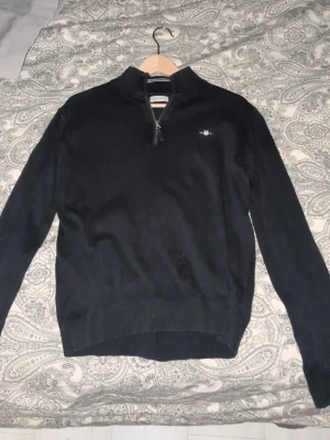 Svart halvzip tröja från Gant - Klassisk marin blå tröja från Gant med halv dragkedja och liten broderad logga på bröstet. Tröjan har hög krage och långa ärmar, perfekt för lager på lager. Tillverkad i mjukt material som känns skönt mot huden.