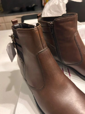 Bruna läder Chelsea boots med dragkedja - Snygga bruna Chelsea boots i slätt läder med dragkedja på sidan och dekorativa spännen vid ankeln. Klassisk siluett med låg klack och elastiska partier för enkel påtagning. Perfekta för dig som vill ha en stilren och tidlös look.