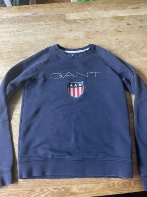 Mörkblå sweatshirt från GANT - Mörkblå sweatshirt från GANT med broderad logga och sköld med stjärnor och ränder på bröstet. Klassisk rund halsringning och långa ärmar. Perfekt för en avslappnad och stilren look.