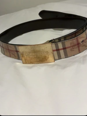 Burberry rutigt bälte med metallspänne - Snyggt bälte från Burberry med klassiskt rutigt mönster i beige, rött och svart. Bältet har ett stort metallspänne med Burberry-logga och insida i mörkbrunt skinn. Perfekt accessoar för att lyfta din outfit.