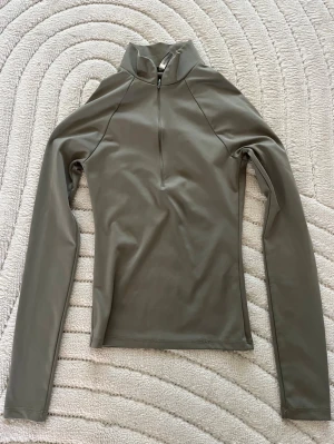 Olivgrön långärmad polotopp - Snygg olivgrön långärmad polotopp med half zip framtill. Tillverkad i ett stretchigt syntetmaterial som sitter tight och ger en sportig vibe. Perfekt att styla med jeans eller träningsbyxor för en clean look. Aldrig använt!!