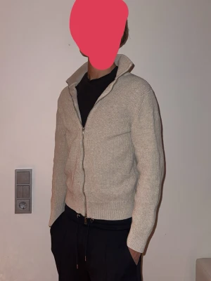 Beige stickad ziptröja med hög krage - Snygg beige stickad tröja med dragkedja framtill och hög krage. Tröjan har en rak passform och ribbade muddar vid ärmslut och nederkant. Perfekt för lager på lager och enkel att matcha med olika outfits.