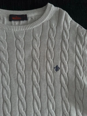 Vit kabelstickad tröja Morris - Kabelstickad vit tröja från Morris med rund halsringning och diskret blå logga broderad på bröstet. Tröjan har långa ärmar och ribbade muddar vid ärmslut och nederkant. Klassisk och stilren design i mjukt material.