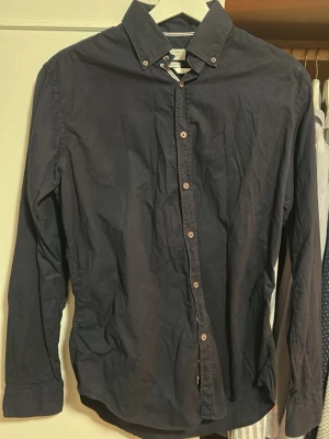Mörkblå skjorta från Gant - Klassisk mörkblå skjorta från Zara med button-down krage och träfärgade knappar. Skjortan har lång ärm och är tillverkad i mjuk bomull, perfekt för en clean och stilren look.