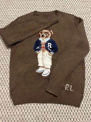 Brun stickad tröja med björnmotiv RL - Stickad brun tröja från Ralph Lauren med ikoniskt björnmotiv på bröstet. Passar ungefär 165 och neråt .Björnen har på sig en blå collegejacka, randig tröja och vita byxor. RL-broderi vid nederkanten. Tröjan har rund hals och ribbade muddar. Perfekt för dig som gillar preppy stil. 