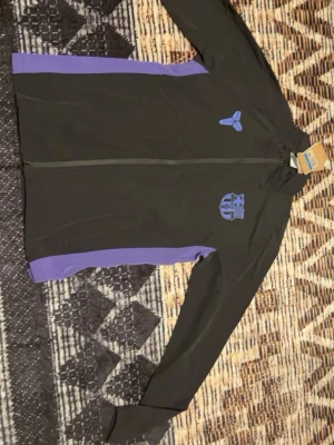 FC Barcelona träningsset svart/lila - Säljer ett FC Barcelona träningsset från Nike i svart med lila detaljer. Setet består av en långärmad zip-jacka och matchande byxor, båda med klubbens och Nikes logga i blått. Perfekt för träning eller uppvärmning. Materialet är lätt och andas.