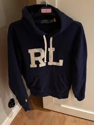 Marinblå hoodie från Polo Ralph Lauren - Snygg marinblå hoodie från Polo Ralph Lauren med stora RL-bokstäver i vitt framtill och klassisk känguruficka. Dragsko i huvan och liten gul logga broderad på ärmen. Perfekt för en avslappnad och trendig look. Inga fläckar eller defekter, som ny.
