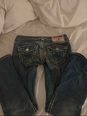Blå True Religion jeans med kontrastsömmar - Säljer ett par blå jeans från True Religion med tydliga vita kontrastsömmar och stora fickor bak med dekorativa knappar. Klassisk femficksmodell i denim med låg midja och raka ben. Märkespatch bak i linningen. Perfekta för en avslappnad streetstil.