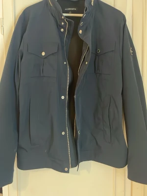 Mörkblå jacka från J.Lindeberg - Snygg mörkblå jacka från J.Lindeberg med silverfärgade knappar och dragkedja. Jackan har två bröstfickor med lock, ståkrage och ett stilrent märke på ärmen. Perfekt för dig som gillar en clean och modern look. Nypris 3500. Helt Ny