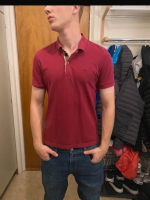 Vinröd pikétröja från Burberry - Säljer en vinröd pikétröja från Burberry med klassisk krage och knappslå med det ikoniska rutmönstret på insidan. Tröjan har korta ärmar och en diskret broderad logga på bröstet. Perfekt för en stilren och avslappnad look.