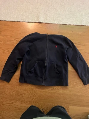 Mörkblå zip hoodie från Polo Ralph Lauren - Snygg mörkblå hoodie med dragkedja från Polo Ralph Lauren. Klassisk design med kängurufickor och den ikoniska röda loggan broderad på bröstet. Perfekt för en chill och stilren look. Tillverkad i mjukt bomullsmaterial.