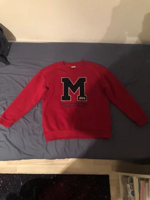 Röd sweatshirt med M-tryck DeFacto - Cool röd sweatshirt från DeFacto med stort svart M-tryck och texten '1986' på bröstet. Tröjan har rund hals, ribbade muddar och är perfekt för en avslappnad stil. Passar dig som gillar collegeinspirerad look.