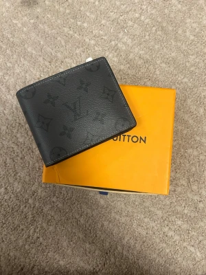 Svart Louis Vuitton plånbok i läder - Snygg svart plånbok från Louis Vuitton i äkta läder med det klassiska monogrammönstret ton-i-ton. Plånboken har en stilren, fyrkantig form och är perfekt för kort och sedlar. Kommer med original orange LV-box.