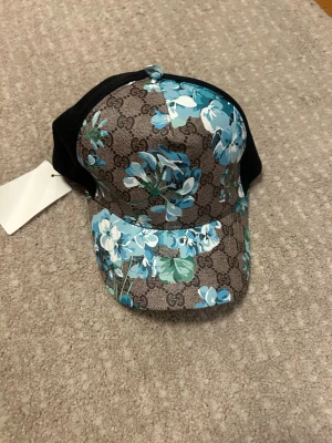 Gucci keps med blå blommor - Snygg keps från Gucci med klassiskt GG-mönster i brunt och beige, dekorerad med blå och vita blommor. Baksidan är i svart mesh för extra ventilation och kepsen har justerbar rem. Perfekt för dig som vill sticka ut med en exklusiv accessoar.