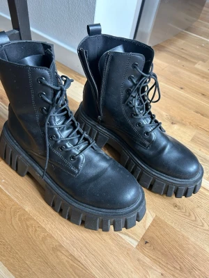 Svarta chunky kängor med grov sula - Säljer ett par svarta kängor med kraftig, grovmönstrad sula och snörning framtill. Kängorna har dragkedja på sidan för enkel på- och avtagning samt dragflik bak. Tillverkade i ett slätt, matt material som ger en cool och edgy look.