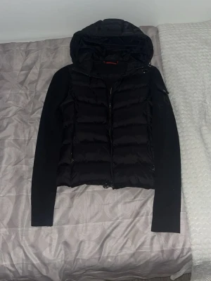 Svart dunväst med huva från Moncler - Snygg svart dunväst från Moncler med stickade ärmar och stor huva. Västen har dragkedja framtill, fickor med dragkedja och en Moncler-logga på ärmen. Perfekt för lager på lager och riktigt trendig look.