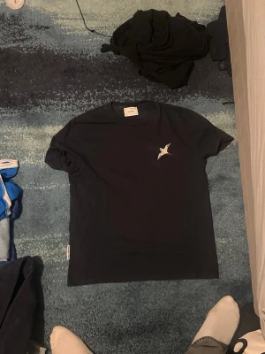 Svart t-shirt med fågeltryck - Snygg svart t-shirt från Axel arigato  med en liten broderad fågel på bröstet. Klassisk passform och rund halsringning. Perfekt till jeans eller shorts för en clean och enkel stil.