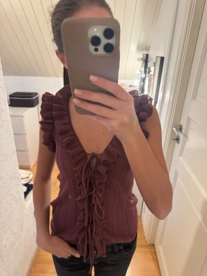 Plommonlila volangtopp med snörning - Supersöt plommonlila topp med volangkant längs v-ringningen och axlarna. Toppen är ärmlös och har snörning framtill. Materialet är ribbat och ger en trendig känsla. 