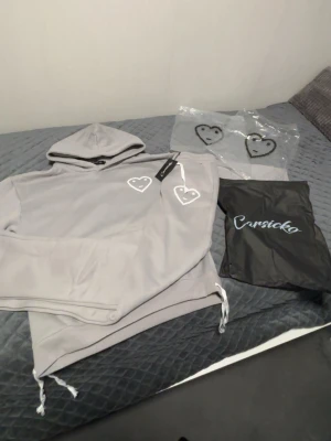 Grå hoodie med hjärtdetaljer Carsicko - Snygg grå hoodie från Carsicko med vita hjärttryck på bröstet och coola snördetaljer i nederkant och huva. Hoodien har en avslappnad passform och är tillverkad i mjukt material, perfekt för en chill streetwear-look.