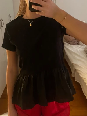 Svart volangblus med kort ärm - Snygg svart blus med mesch volang nedtill och korta ärmar. Modellen har en rund halsringning och en lös passform som ger en avslappnad men trendig look. Perfekt att matcha med jeans eller kjol för en cool stil.
