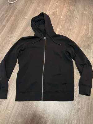 Svart zip hoodie med huva från Jack & Jones - Klassisk svart hoodie med dragkedja framtill och stor huva med dragsko. Hoodien har två rymliga fickor på magen och ribbade muddar vid ärmar och nederkant. Perfekt för en avslappnad och stilren look. Från Jack & Jones