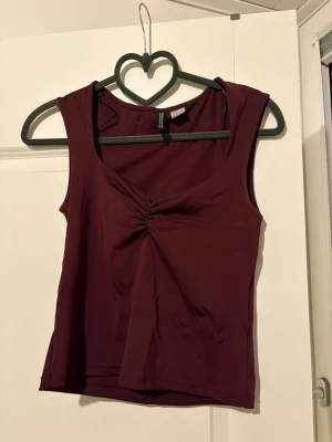 Vinröd ärmlös topp från Divided - Snygg vinröd ärmlös topp från Divided med twistad detalj framtill vid bysten. Toppen är croppad och har en mjuk, stretchig känsla i bomullsmix. 