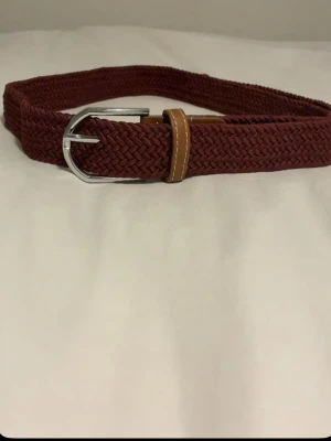 Vinrött flätat bälte med skinn-detalj - Snyggt vinrött flätat bälte med silverfärgat spänne och bruna skinn-detaljer. Perfekt accessoar för att lyfta din outfit med en trendig touch. Bältet har en flexibel design och passar till många olika stilar.