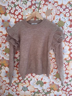 Beige stickad tröja med volangärmar - Supersöt beige stickad tröja med volanger på axlarna och långa ärmar. Tröjan har rund halsringning och ribbade muddar vid ärmslut och nederkant. Perfekt för dig som vill ha en trendig och mysig look.