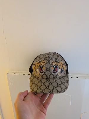 "ÄKTA" Gucci keps med tigertryck - Säljer en snygg Gucci keps med klassiskt GG-mönster i beige och brunt. Framtill finns två detaljerade tigerhuvuden i gult och svart. Baksidan är i svart mesh för extra ventilation. Perfekt för dig som vill sticka ut med en exklusiv accessoar.