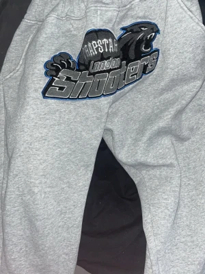 Gråa Trapstar mjukisbyxor L - Säljer ett par gråa mjukisbyxor från Trapstar med broderad London Shooters-logga och pantermotiv framtill. Byxorna har resår i midjan och är gjorda i mjukt sweatshirtmaterial, perfekta för en chill och avslappnad stil.