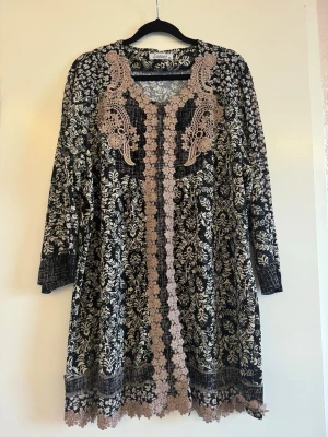 Svart blommig klänning med spets - Unik svart klänning från Daddato med blommigt mönster i beige och dekorativa spetsdetaljer framtill. Klänningen har lång ärm, v-ringning och fina broderade inslag som ger en bohemisk känsla. Perfekt för dig som gillar detaljer och mönster. Den är köpt utomlands. Skulle säga att storleken är L då den sitter perfekt på min mamma som har storlek L. Den har en loose passform alltså sitter inte tajt. Bröstmåttet är ca 110cm (55cm x 2).