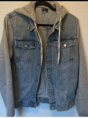 Jeansjacka med grå hoodie från Divided - Snygg ljusblå jeansjacka från Divided med grå sweatshirt-ärmar och huva. Jackan har två bröstfickor med knappar och dragsko i huvan för en avslappnad streetstil. Perfekt för dig som gillar lager på lager och vill ha en cool vardagslook.