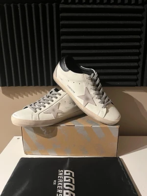 Golden goose skor  - Snygga vita sneakers från Golden Goose med grå mockastjärna på sidan, grå skosnören och svart hälparti. Jätte bra skick och kan diskuter om pris. De funkar även för folk som har storlek 44