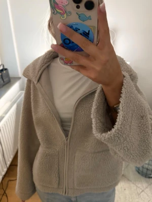 Beige teddyjacka med dragkedja - Mysig beige teddyjacka med dragkedja framtill och stora fickor. Jackan har en bred krage och är tillverkad i fluffigt teddy-material som ger en avslappnad och trendig look. Perfekt för kyliga dagar och enkel att matcha med jeans eller leggings.